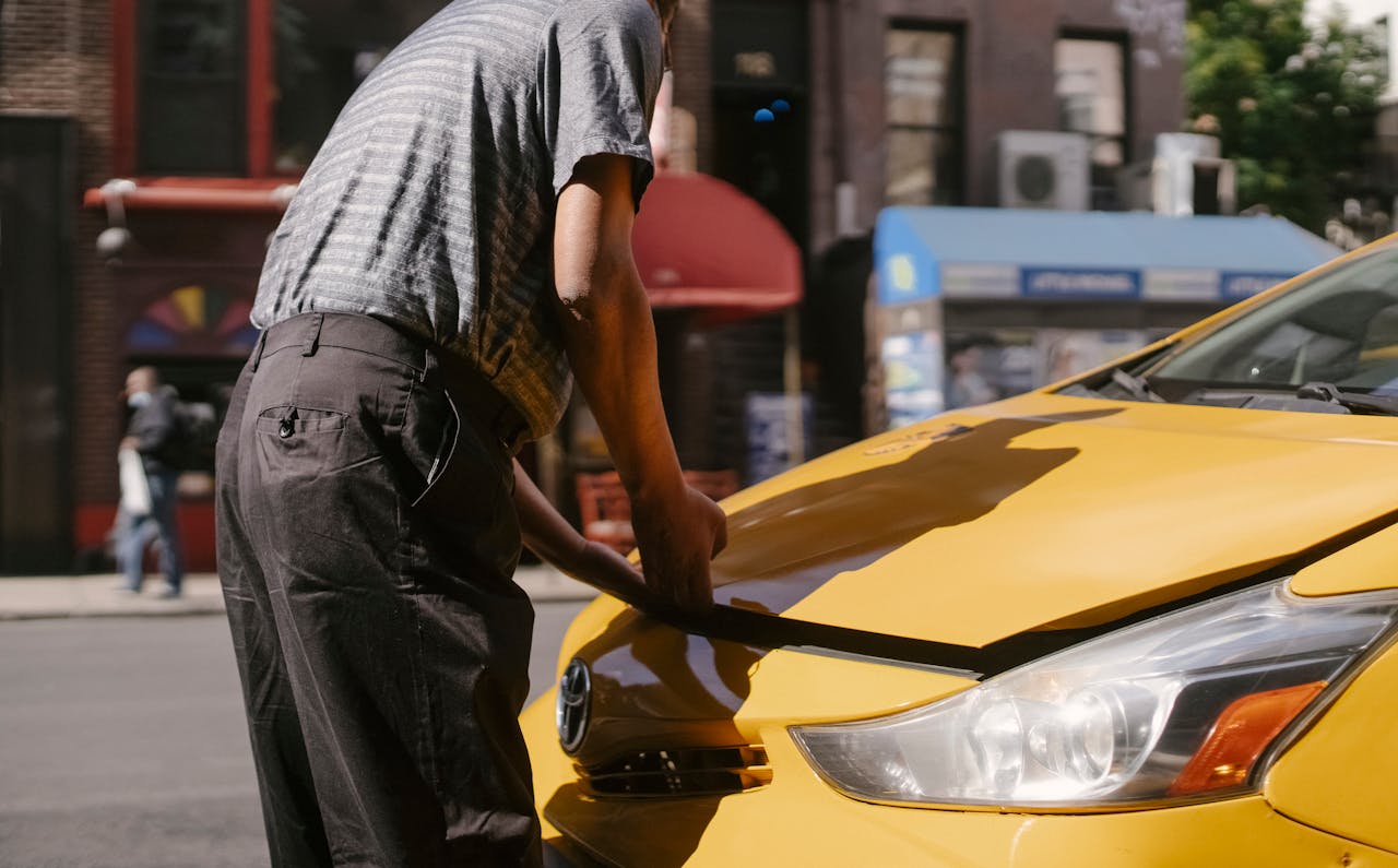 Man inspecting open hood of a yellow taxi on city street.